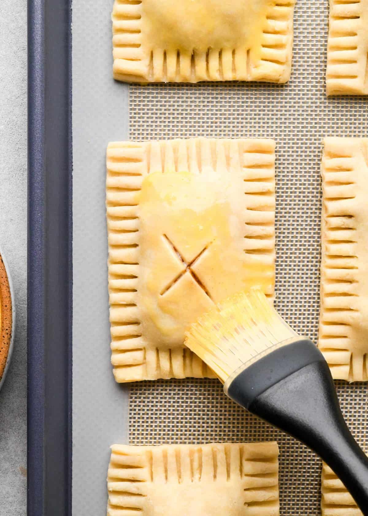 a pastry brush brushing the egg wash on the top of an apple hand pie on a baking sheet