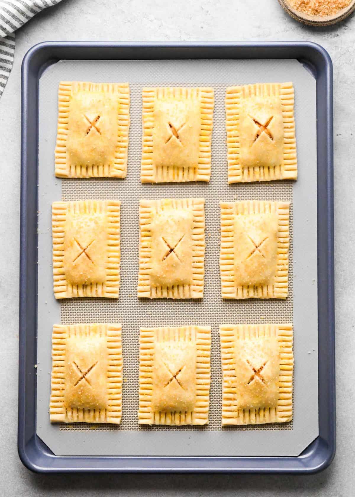 9 assembled apple hand pies on a parchment lined baking sheet with an "X" cut into the center of each one brushed with egg wash before baking