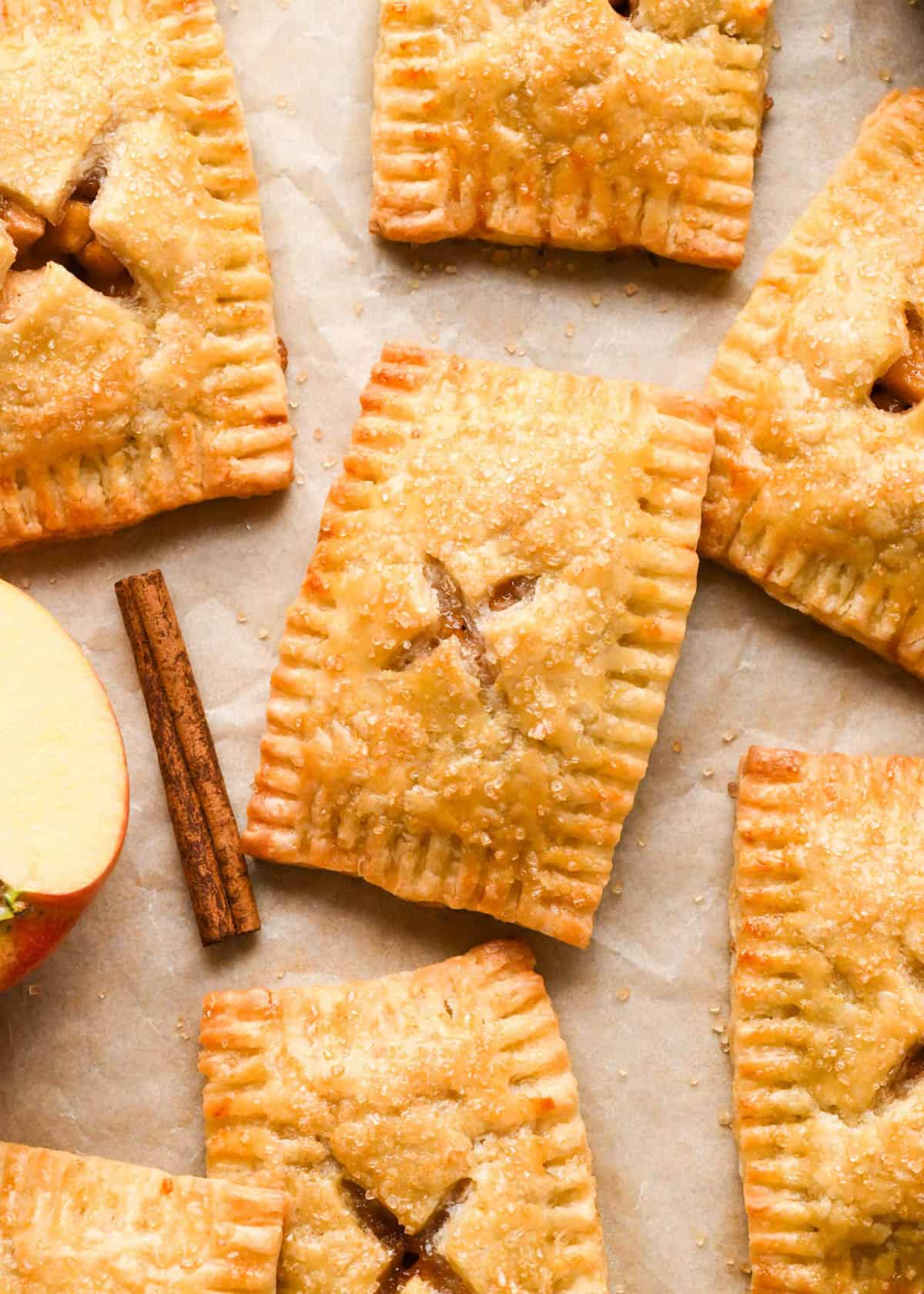 6 an apple hand pies with crimped edges, and cinnamon apple filling peeking through vented tops with an "X" cut into them, sprinkled with raw sugar on parchment paper surrounded by cinnamon sticks and apples