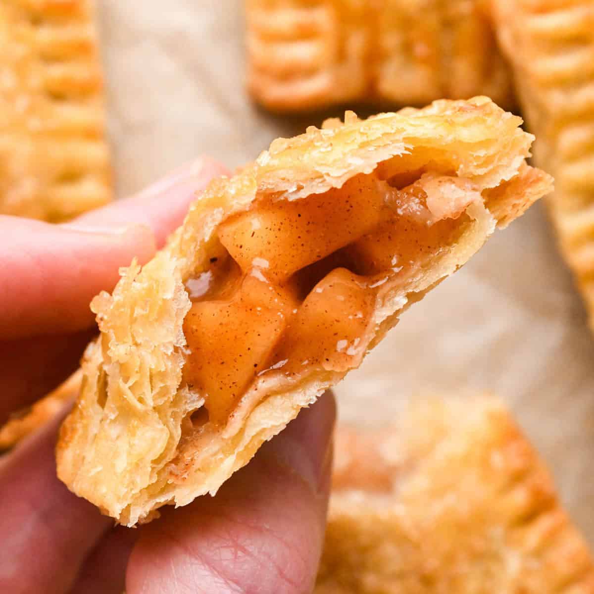 a hand holding a half of an apple hand pie so the gooey cinnamon apple filling is showing with more apple hand pies in the backgorund