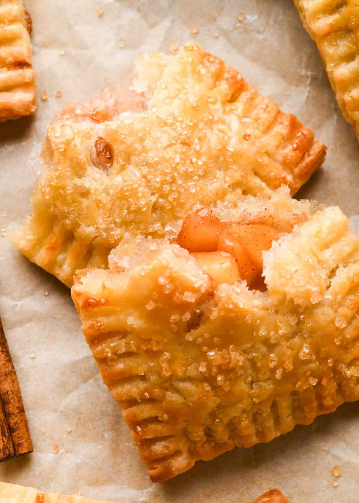 apple hand pie cut in half with raw sugar sprinkled on top, on parchment paper with other hand pies and a cinnamon stick around it