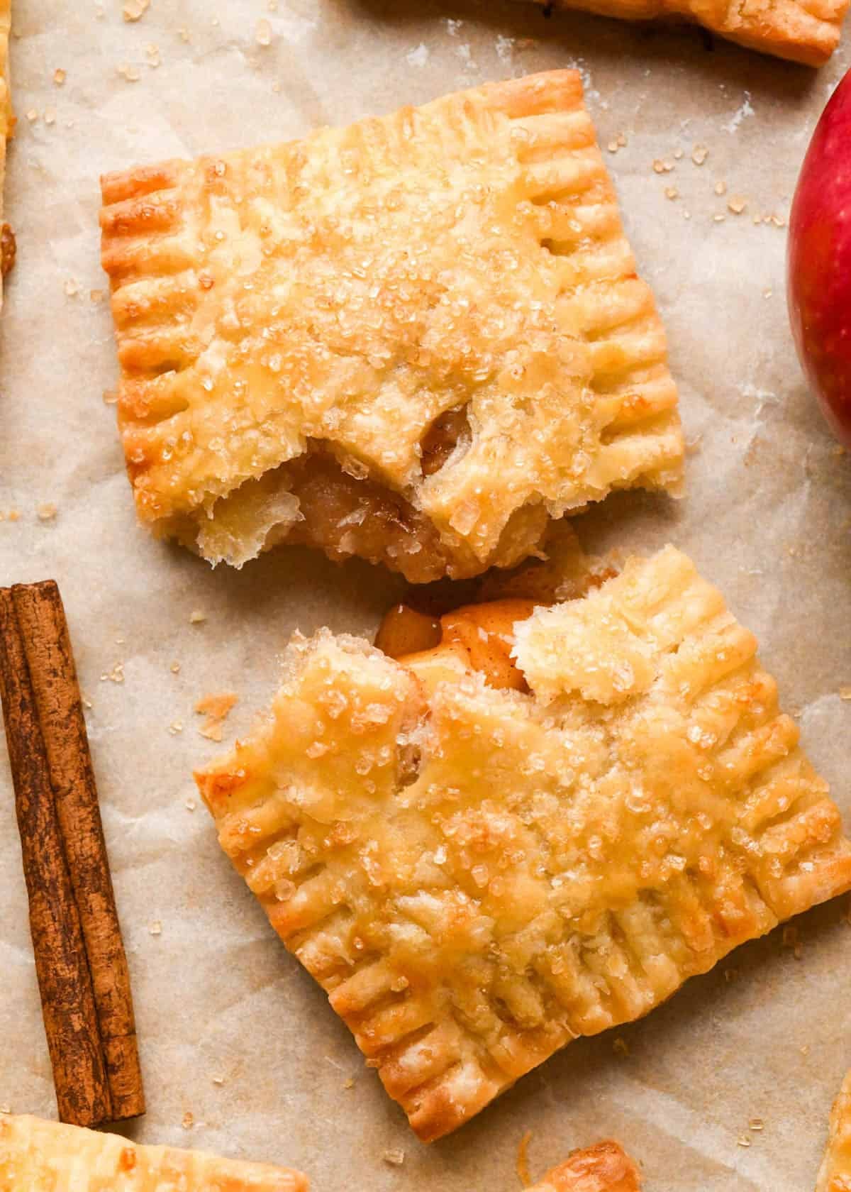 an apple hand pie torn in half with crimped edges, and cinnamon apple filling peeking through a vented top with an "X" cut into it, sprinkled with raw sugar on parchment paper surrounded by cinnamon sticks, apples and two other apple pie hand pies