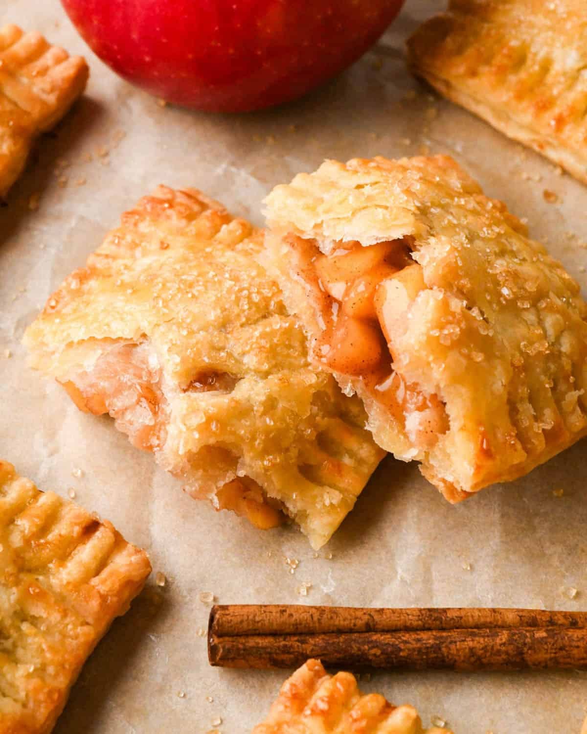 apple hand pie cut in half with raw sugar sprinkled on top, the apple filling is visible, on parchment paper with other hand pies, an apple, and a cinnamon stick around it