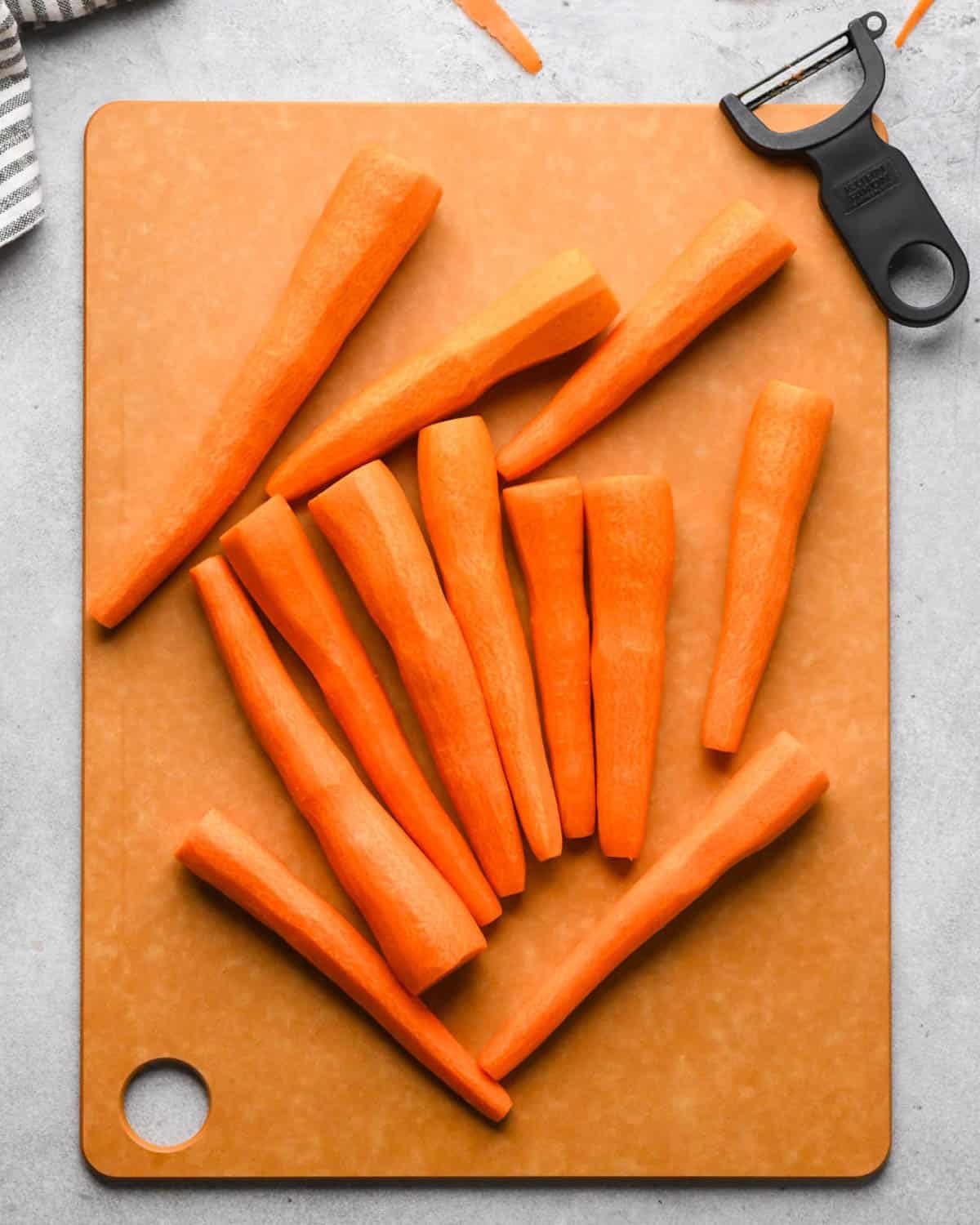 making glazed maple carrots - whole carrots peeled on a brown cutting board with the peeler in the top right corner