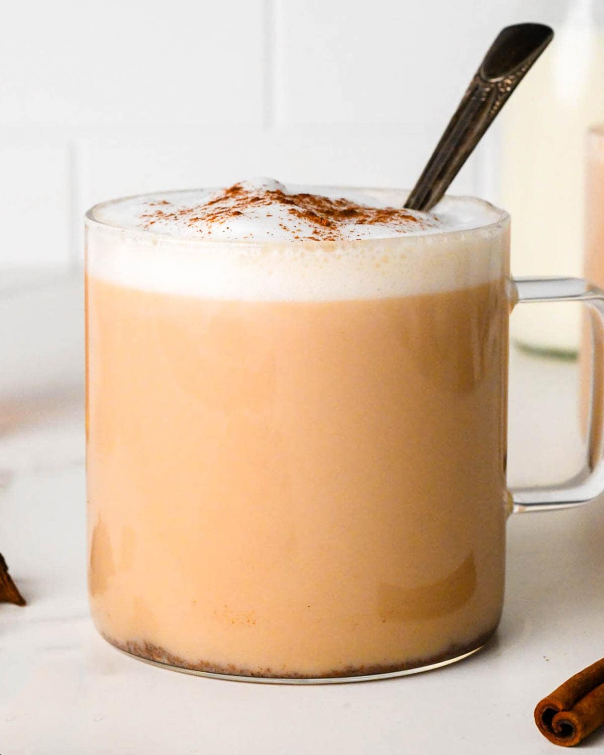 chai latte in a clear glass mug topped with foamy milk and a sprinkle of cinnamon with a metal spoon inside, milk, and another chai tea are in the background
