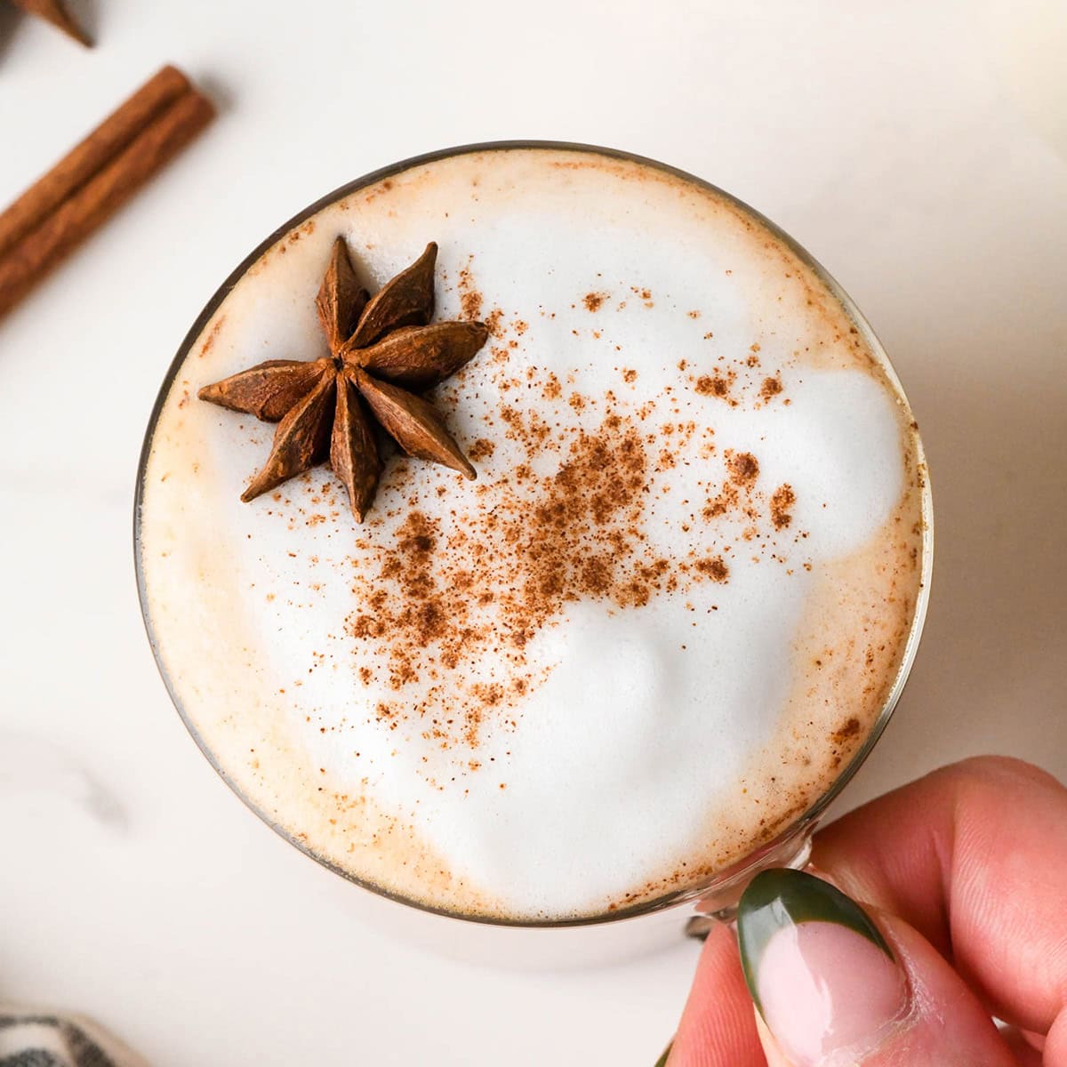 overhead photo of a hand holding a chai latte in a glass mug topped with foamy milk, a sprinkle of cinnamon and star anise, with cinnamon sticks in the background