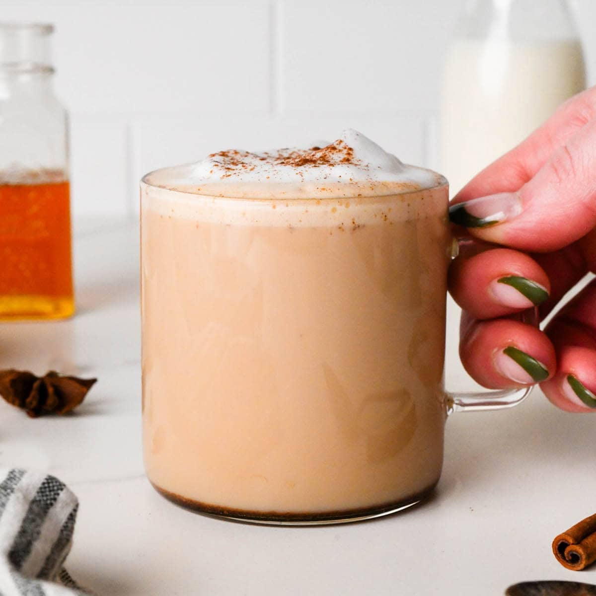 a hand holding a chai latte in a clear glass mug topped with foamy milk and a sprinkle of cinnamon