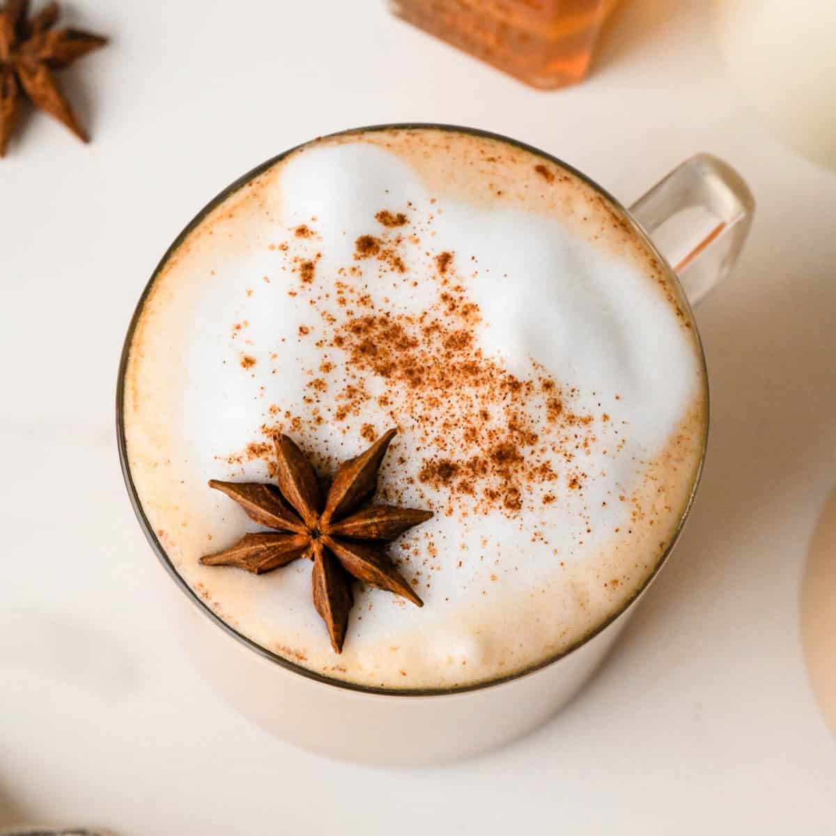 overhead photo of a chai latte in a glass mug topped with foamy milk, a sprinkle of cinnamon and star anise