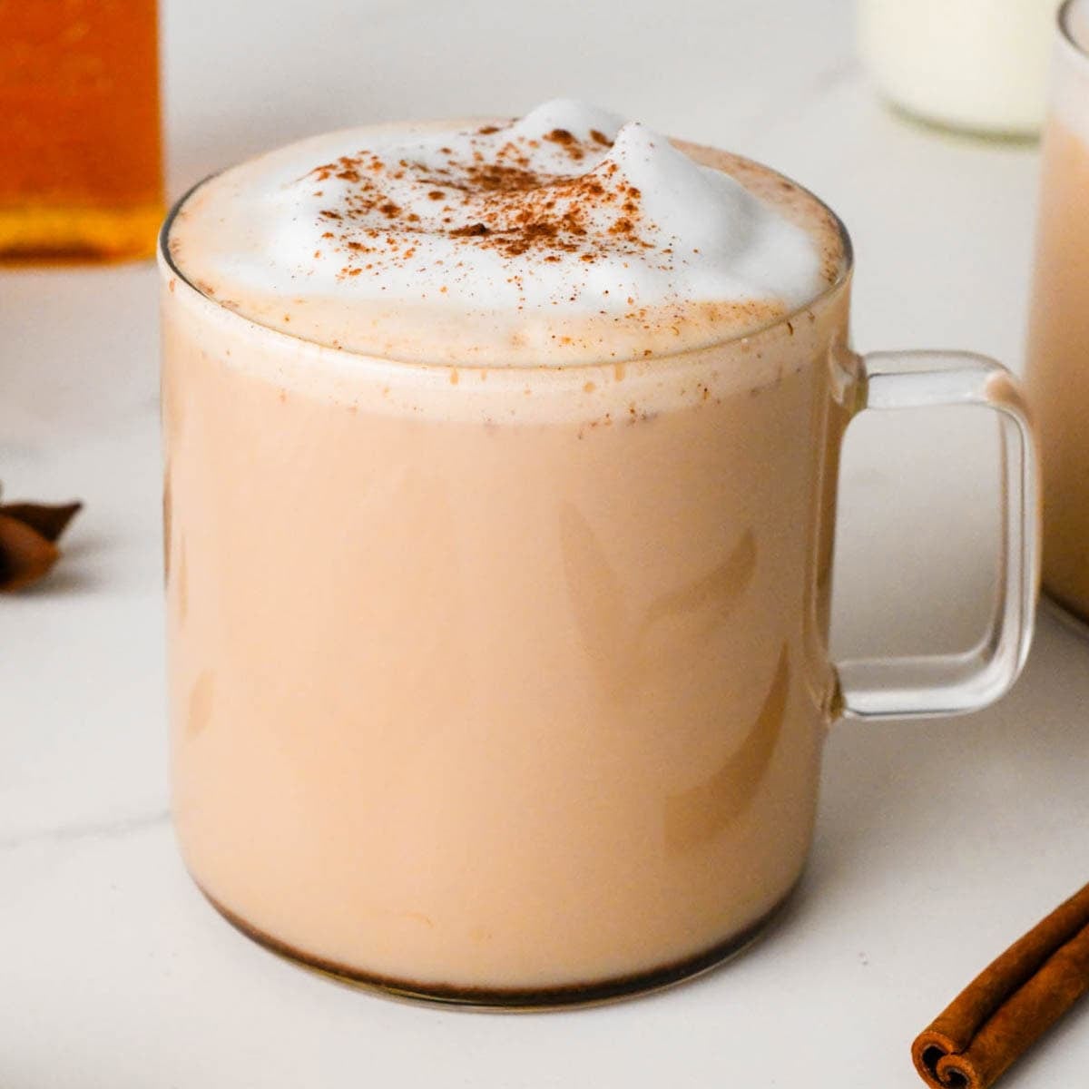 chai latte in a clear glass mug topped with foamy milk and a sprinkle of cinnamon, brewed tea, milk, and another chai tea are in the background