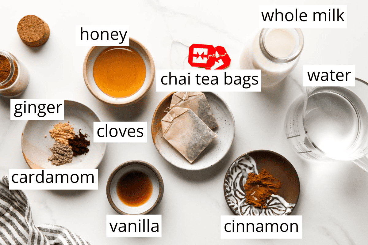 overhead photo of the ingredients for this chai latte recipe in bowls and labeled like honey, tea bags, water, milk, and spices