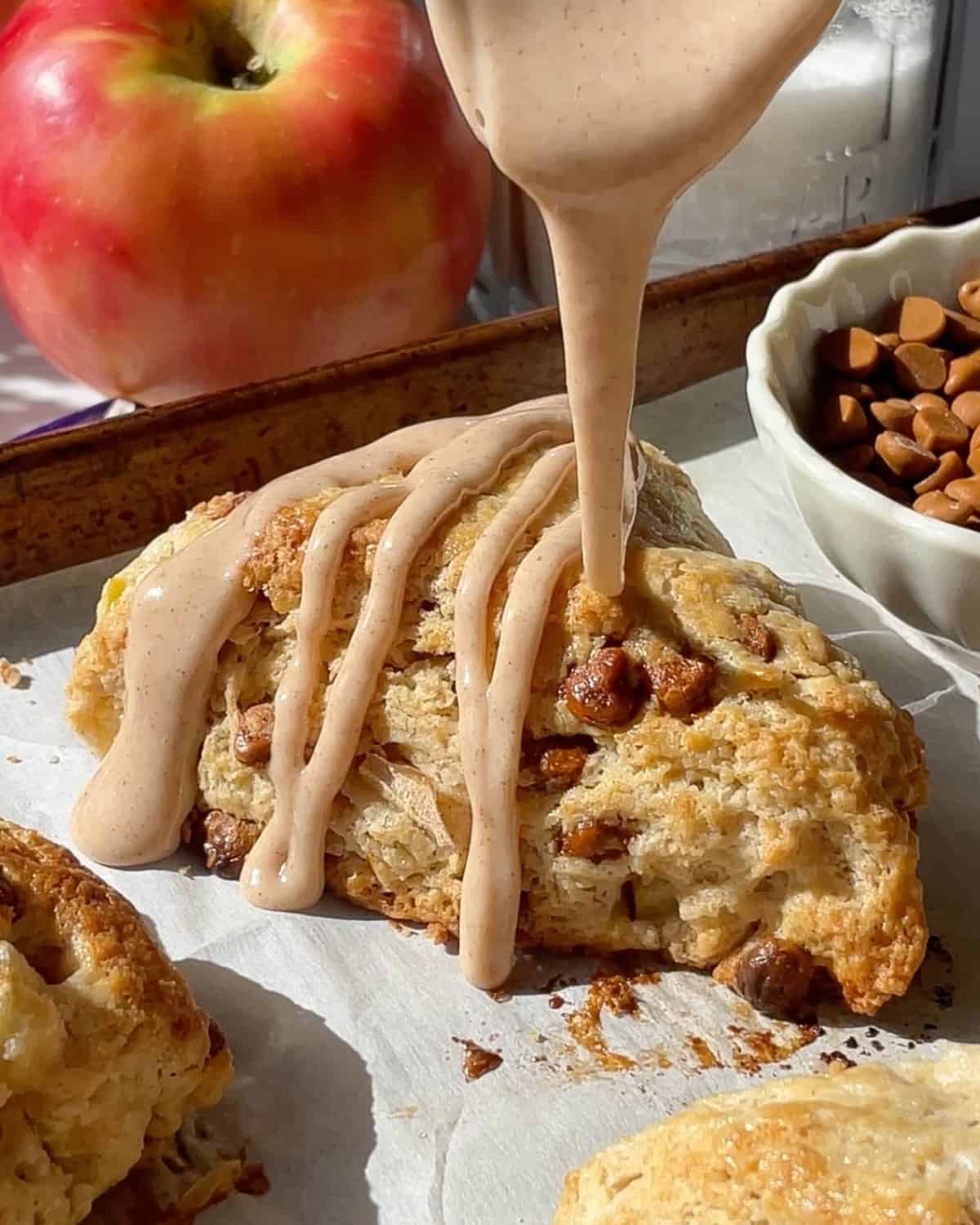 cinnamon glaze being drizzled over an apple scone on a baking sheet lined with parchment paper with a silver spoon, there are more scones and apples in around it