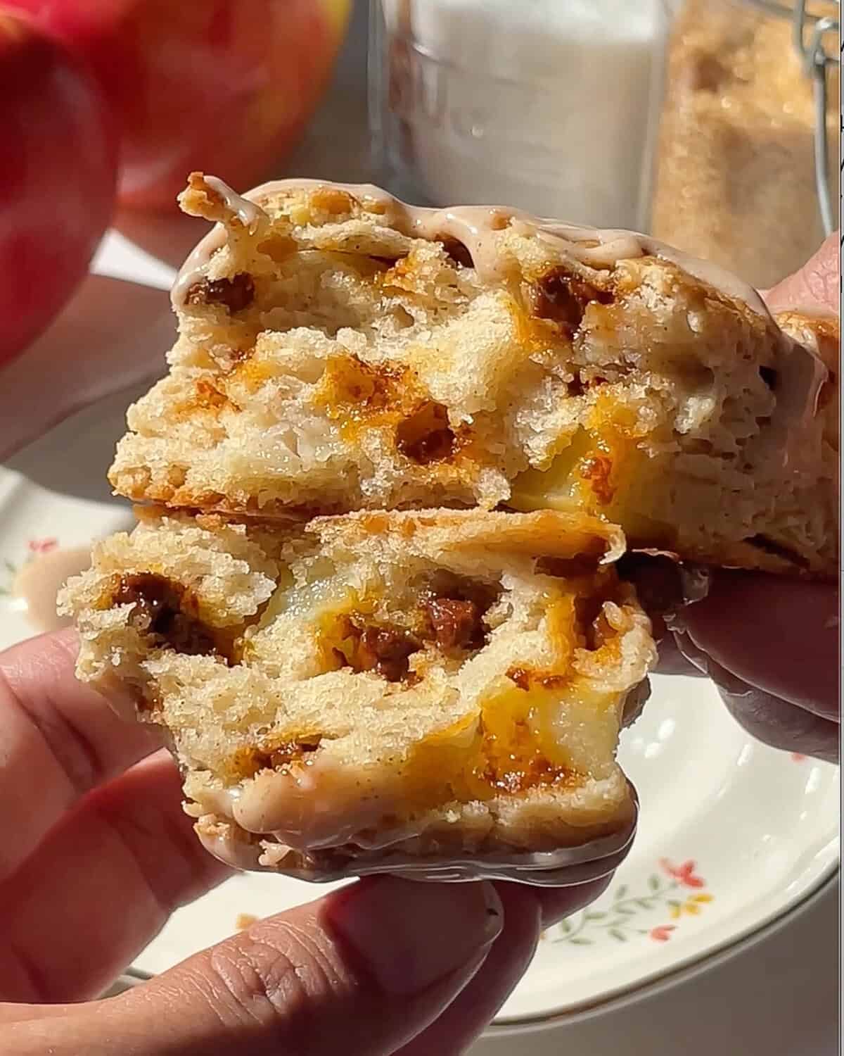a hand holding two halves of a cinnamon apple scone with cinnamon glaze stacked on each other so the chunks of apples, moist interior and cinnamon chips are visible. There are apples and sugar in the background