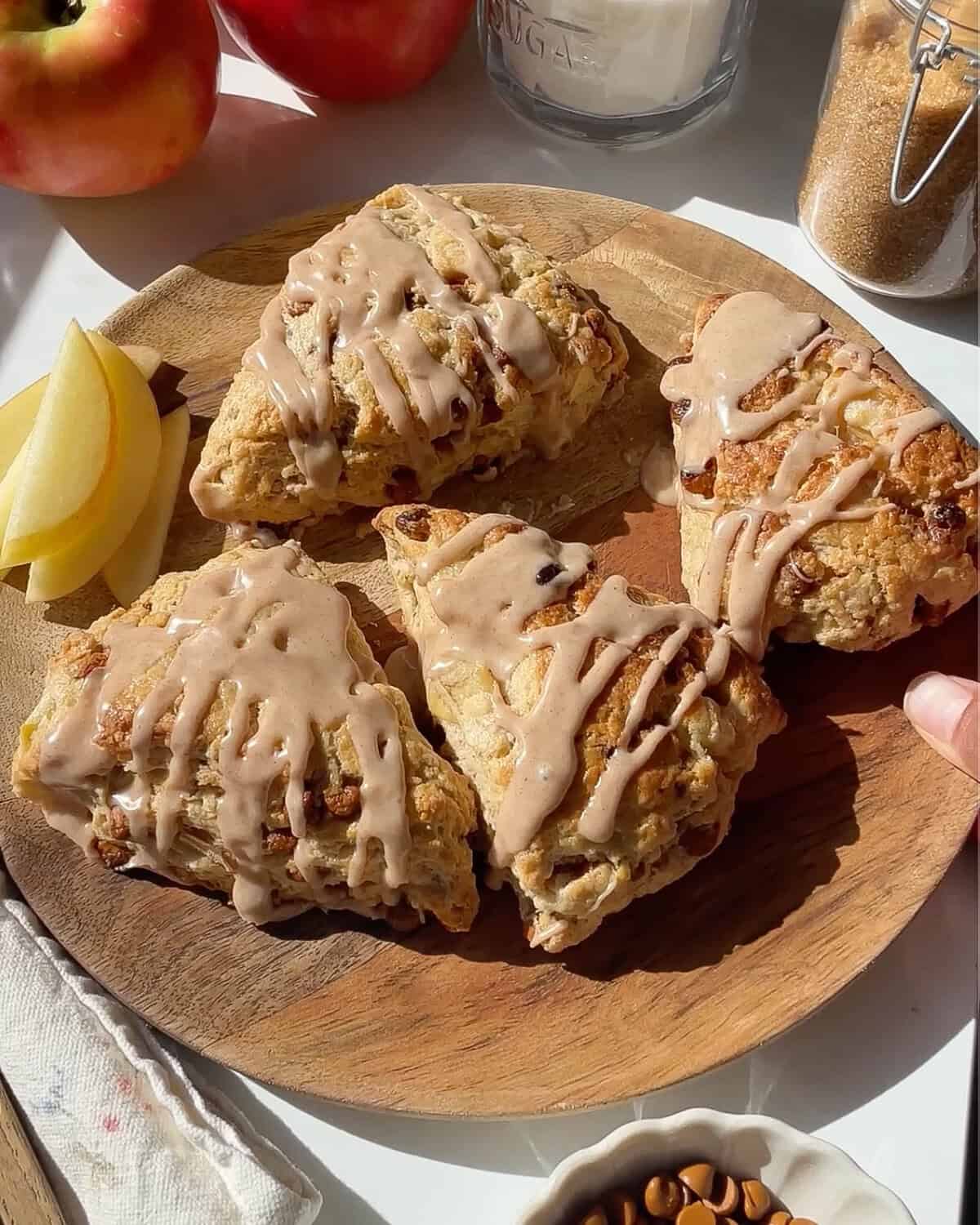4 cinnamon apple scones with cinnamon glaze on a round wooden plate with sliced apples on the plate. there are apples and sugar in the background