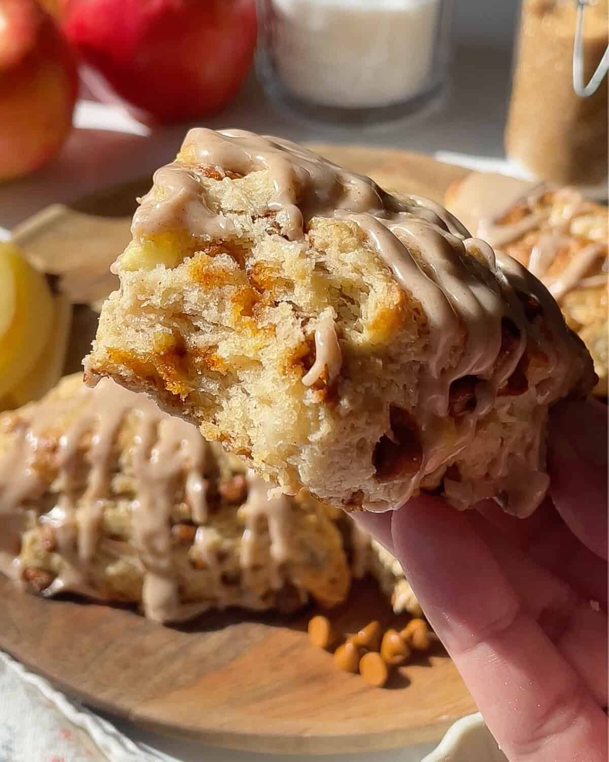 d holding an apple cinnamon scone with cinnamon glaze with a bite taken out of it so the moist texture can be seen over a plate of apple scones with apples in the background.