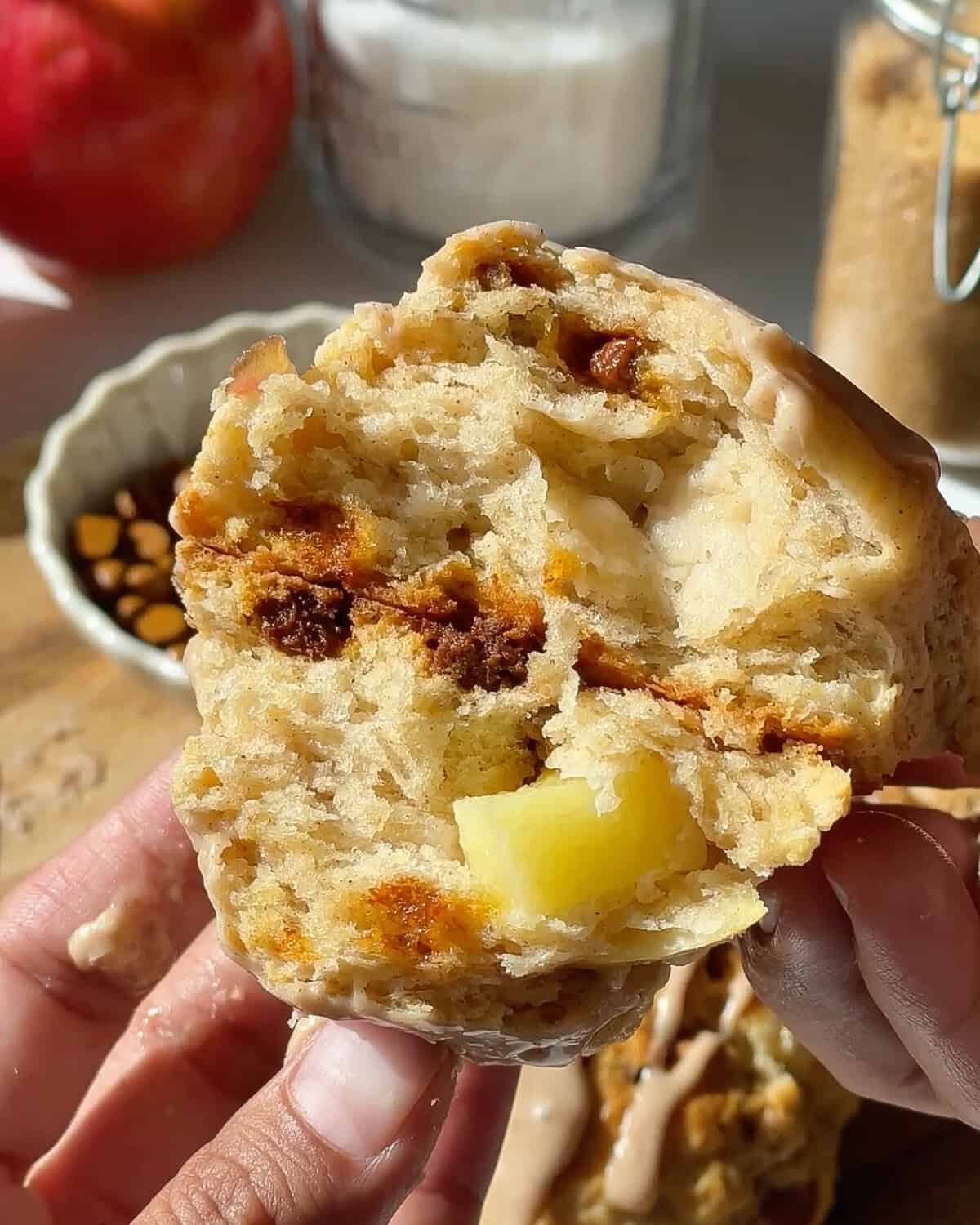 a hand holding two halves of a cinnamon apple scone stacked on each other so the chunks of apples, moist interior and cinnamon chips are visible. There is an apple and bowl of cinnamon chips in the background.