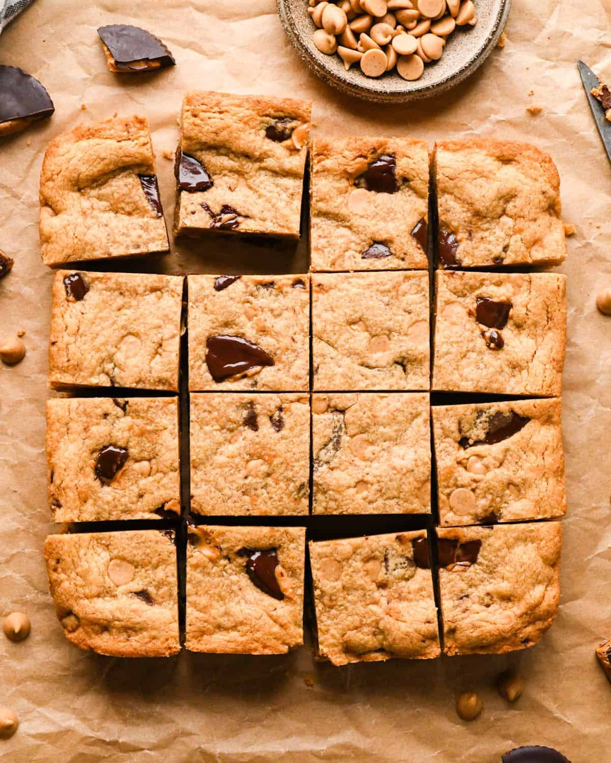 peanut butter blondies on parchment paper cut into 16 squares surrounded by peanut butter chips and peanut butter cups