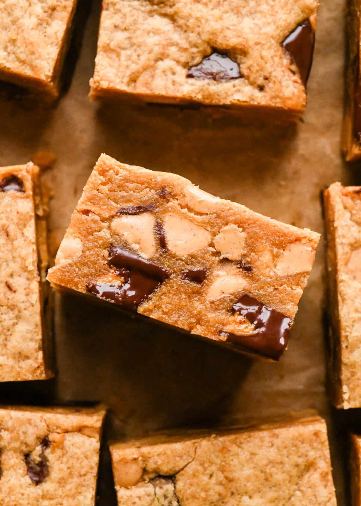 1 peanut butter blondie turned on its side so the fudgy texture is visible along with with melty peanut butter cups and peanut butter chips. The blondes are on parchment paper and surrounded by more peanut butter blondies