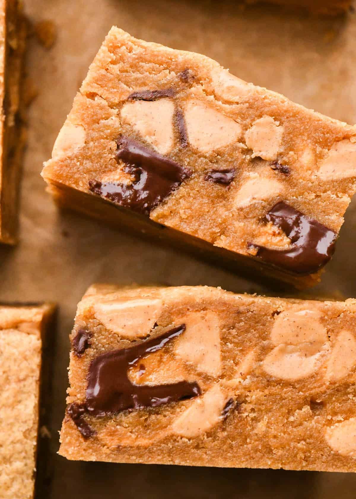 2 peanut butter blondies turned on their side so the fudgy texture is visible along with with melty peanut butter cups and peanut butter chips. The blondes are on parchment paper and surrounded by more peanut butter blondies