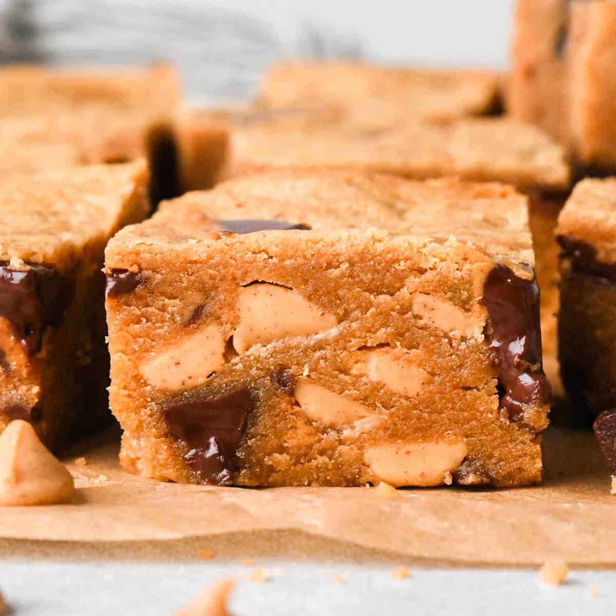 front view of a peanut butter blondie, the fudgy texture is visible along with with melty peanut butter cups and peanut butter chips. The blonde is on parchment paper and surrounded by more peanut butter blondies, peanut butter chips and peanut butter cups