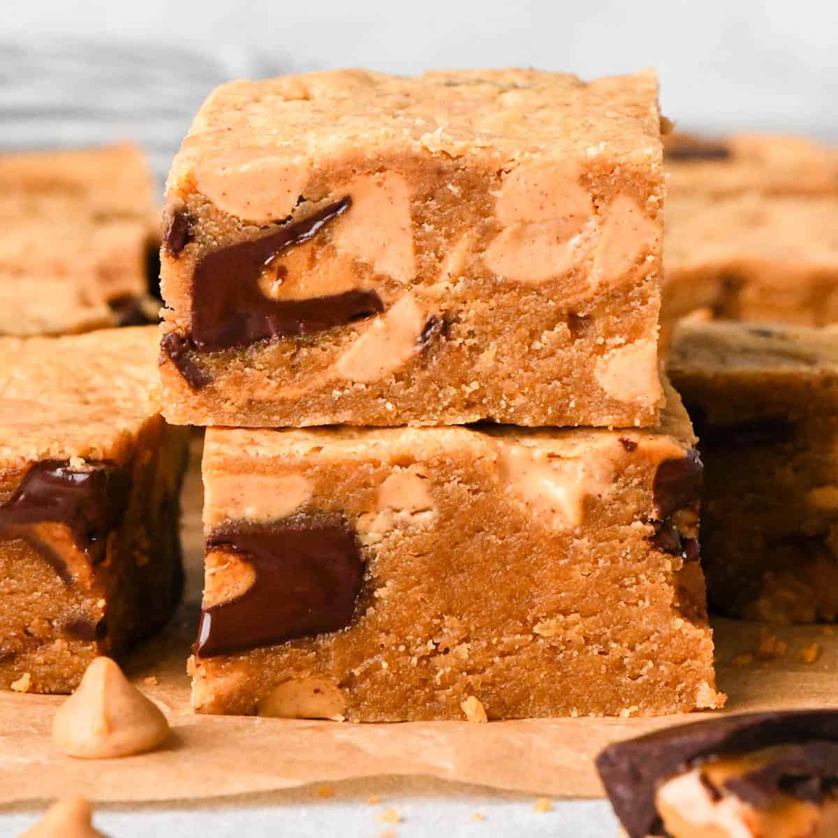 a stack of 2 peanut butter blondies, the fudgy texture is visible and studded with melty peanut butter cups and peanut butter chips. The stack is on parchment paper and surrounded by more peanut butter blondies, peanut butter chips and peanut butter cups
