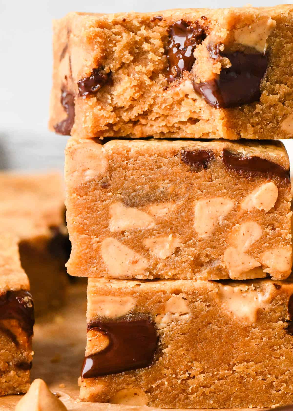 close up photo of a stack of 3 peanut butter blondies, the top one has a bite taken out of it, in all of them the fudgy texture is visible and studded with melty peanut butter cups and peanut butter chips. The stack is on parchment paper and surrounded by more peanut butter blondies, peanut butter chips and peanut butter cups