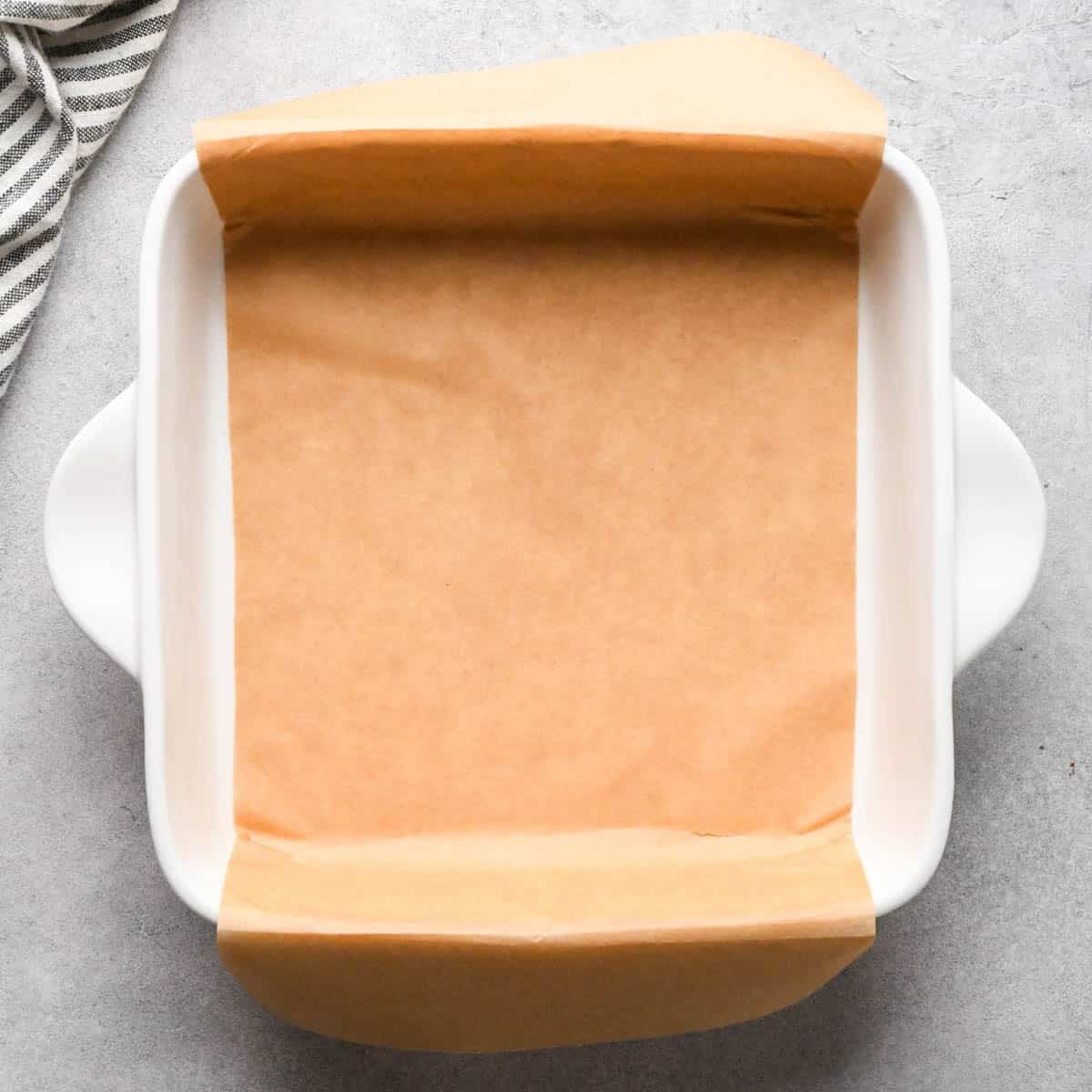 making peanut butter blondies - a white ceramic baking dish lined with brown parchment paper, there's a striped towel in the upper left corner