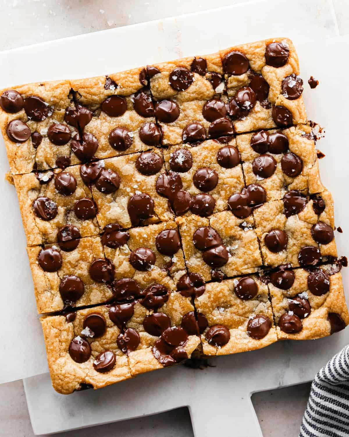 overhead view of brown butter cookie bars on a marble cutting board cut into 16 squares with melty chocolate chips and topped with flaky sea salt.