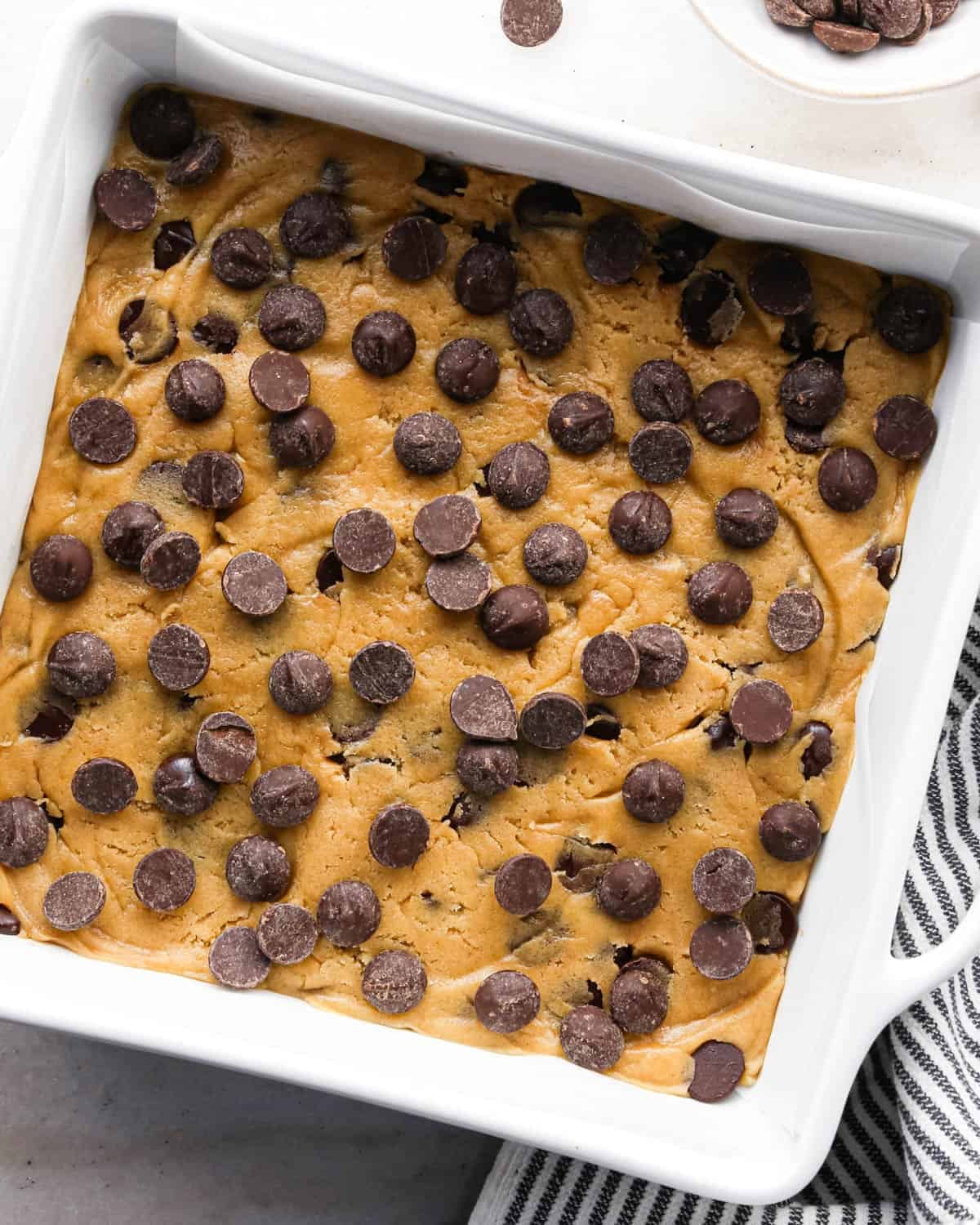 making brown butter cookie bars - dough pressed into a parchment-lined white square baking dish and topped with chocolate chips