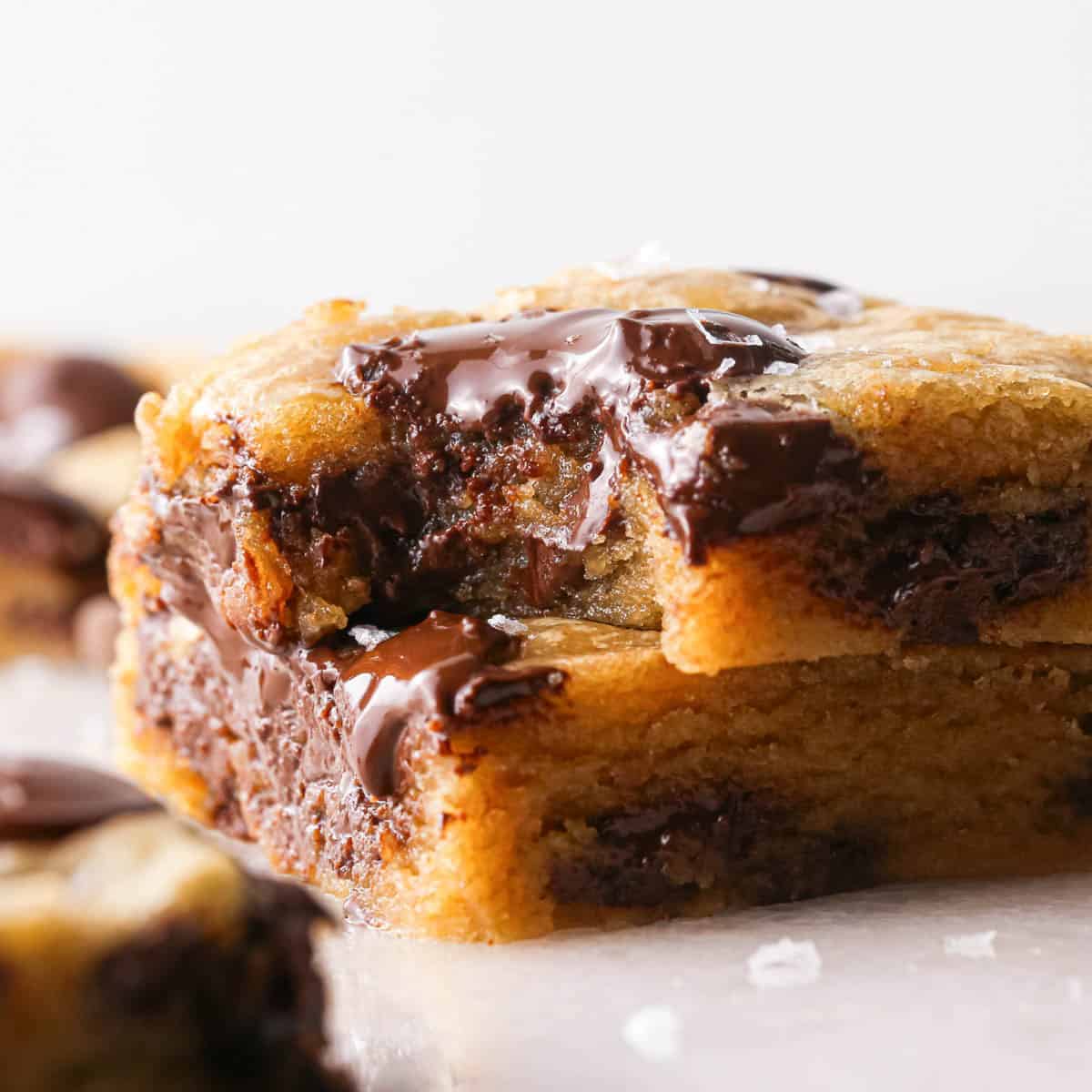 a stack of 2 brown butter chocolate chip cookie bars with melty chocolate chips and a gooey texture, topped with flaky sea salt. The top one has a bite taken out of it. The stack is surrounded by more cookie bars and flaky sea salt