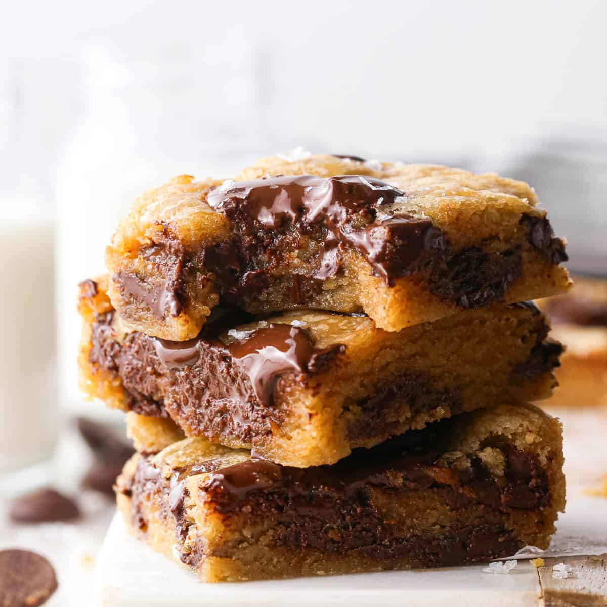a stack of 3 brown butter chocolate chip cookie bars with melty chocolate chips and a gooey texture, topped with flaky sea salt. The top one has a bite taken out of it. The stack is on parchment paper and surrounded by more cookie bars and chocolate chips