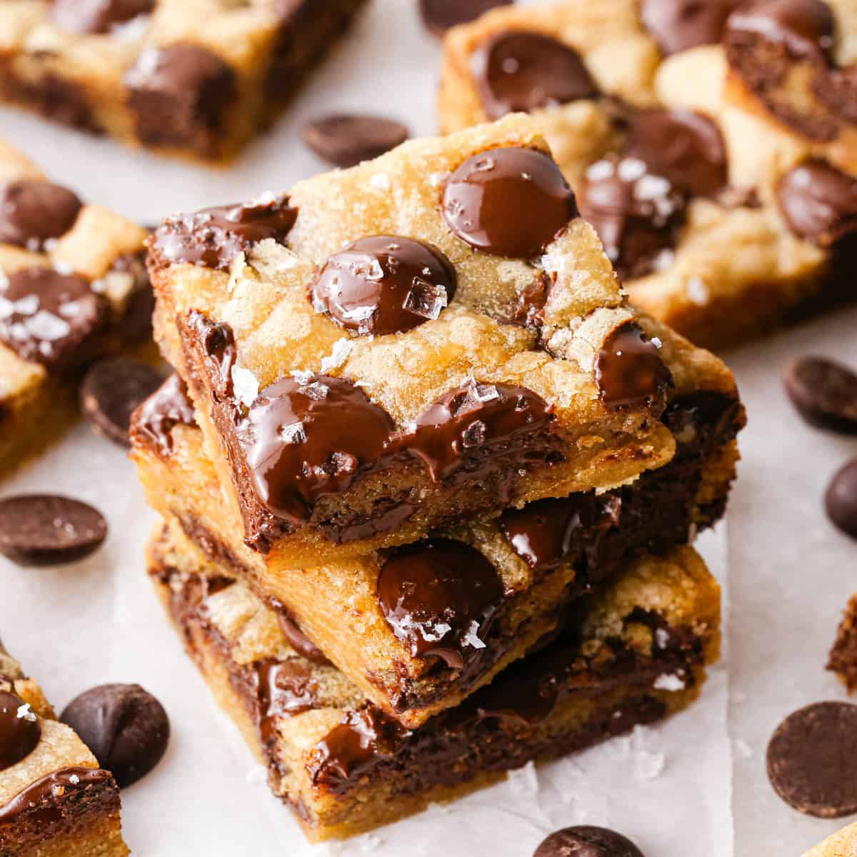 a stack of 3 brown butter chocolate chip cookie bars with melty chocolate chips and a gooey texture, topped with flaky sea salt. The stack is on parchment paper and surrounded by more cookie bars and chocolate chips