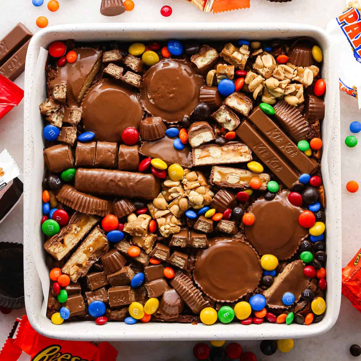overhead photo of leftover halloween candy brownies topped with tons of candy like twix, peanut butter cups, and M&Ms, in a white, parchment-lined square baking dish, surrounded by more candy and wrappers
