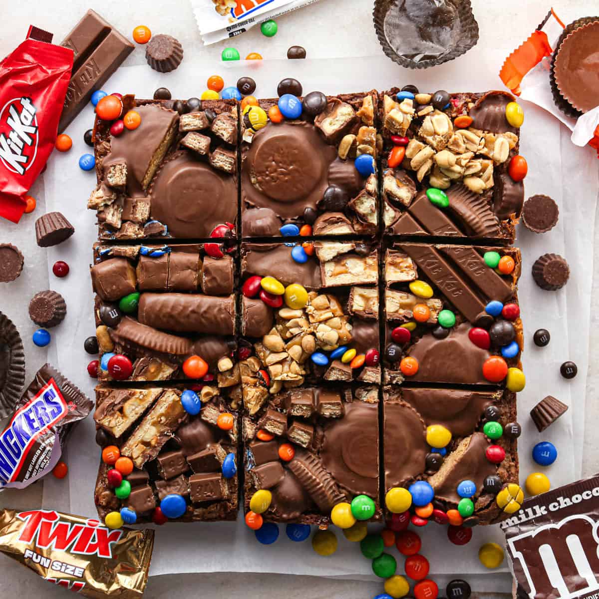 overhead photo of leftover halloween candy brownies topped with tons of candy like twix, peanut butter cups, and M&Ms, cut into 9 squares on parchment paper, surrounded by more candy and wrappers