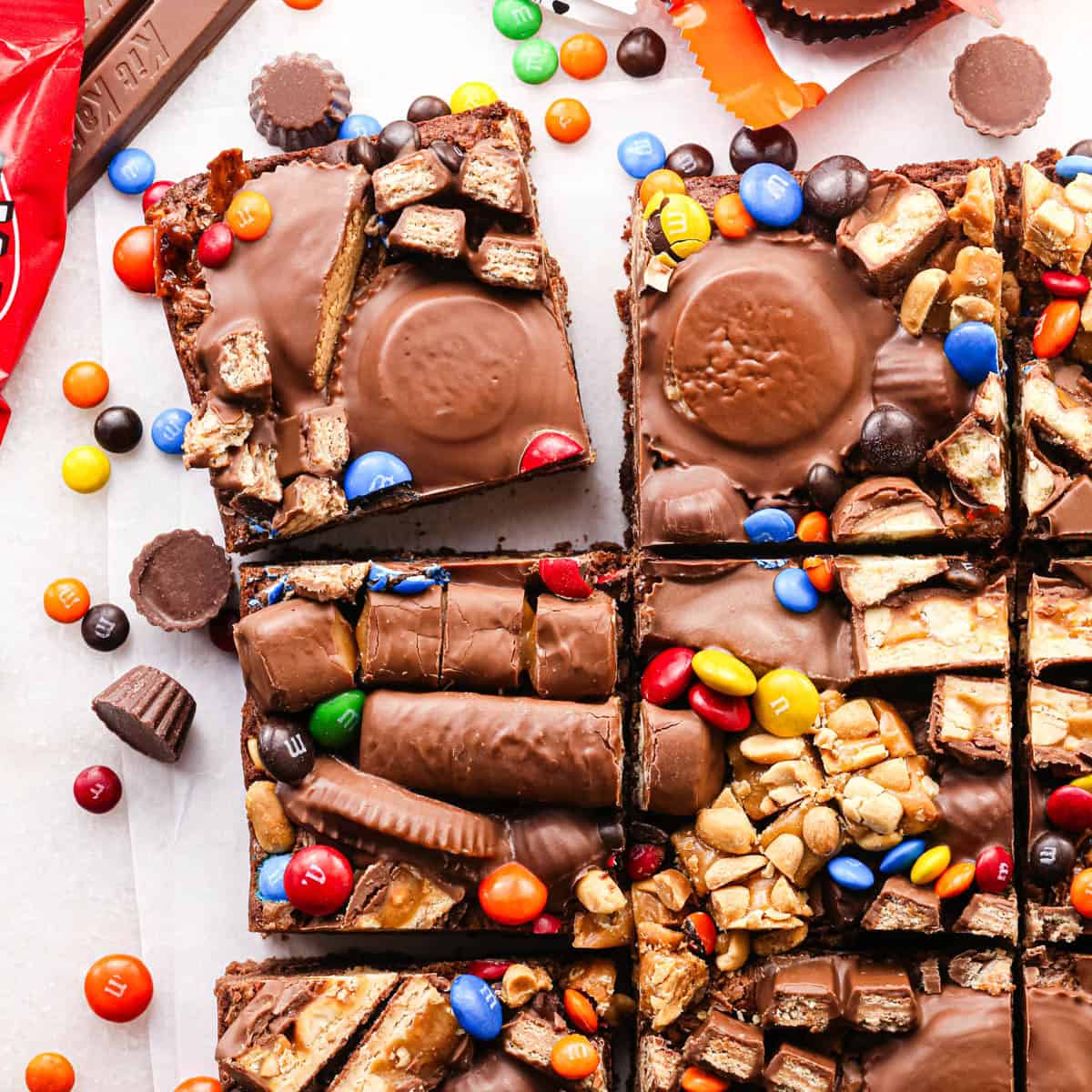 overhead photo of leftover halloween candy brownies topped with tons of candy like twix, peanut butter cups, and M&Ms, cut into squares on parchment paper, surrounded by more candy and wrappers