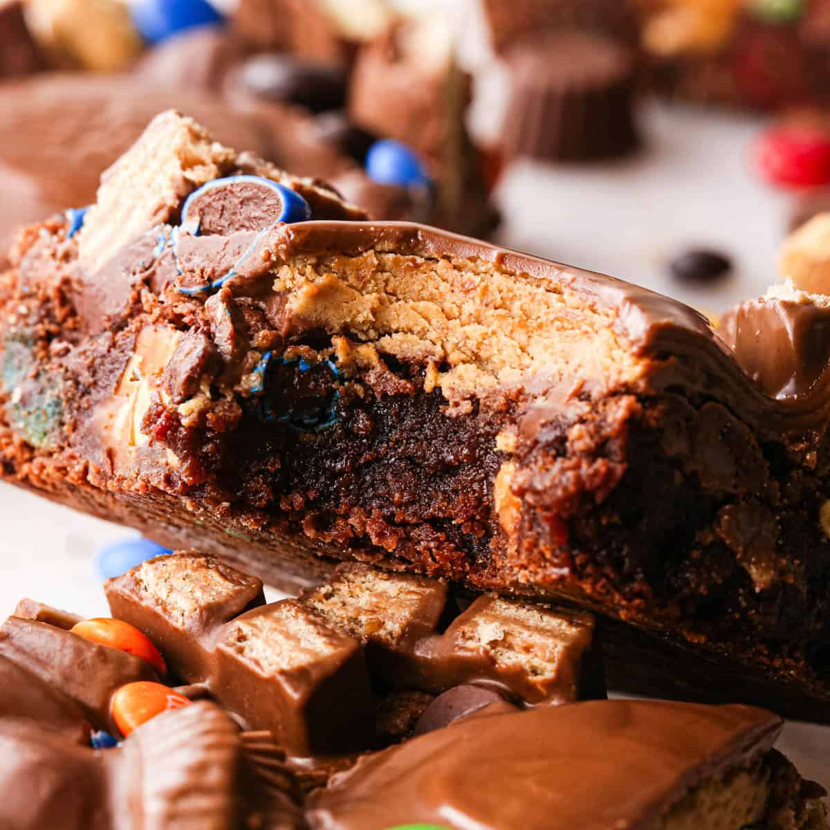 close up photo of a halloween brownie with a bite taken out of it so the fudgy texture and candy inside is visible. It's resting on another brownie with more halloween brownies and candy around it