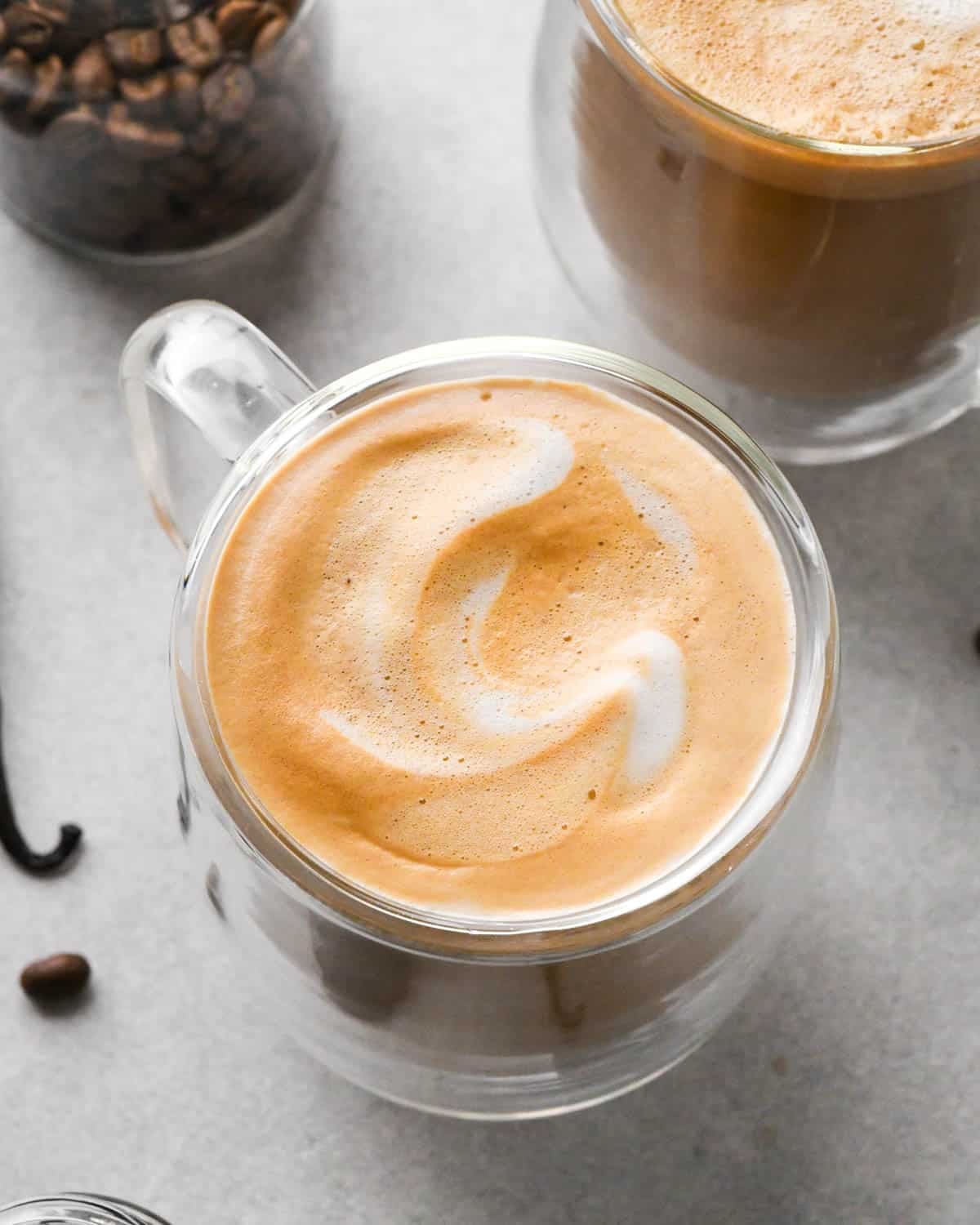 overhead view of a hot vanilla latte in a glass mug with the foamy milk on top visible. There's another latte, vanilla bean, coffee bean and a jar of coffee beans in the background