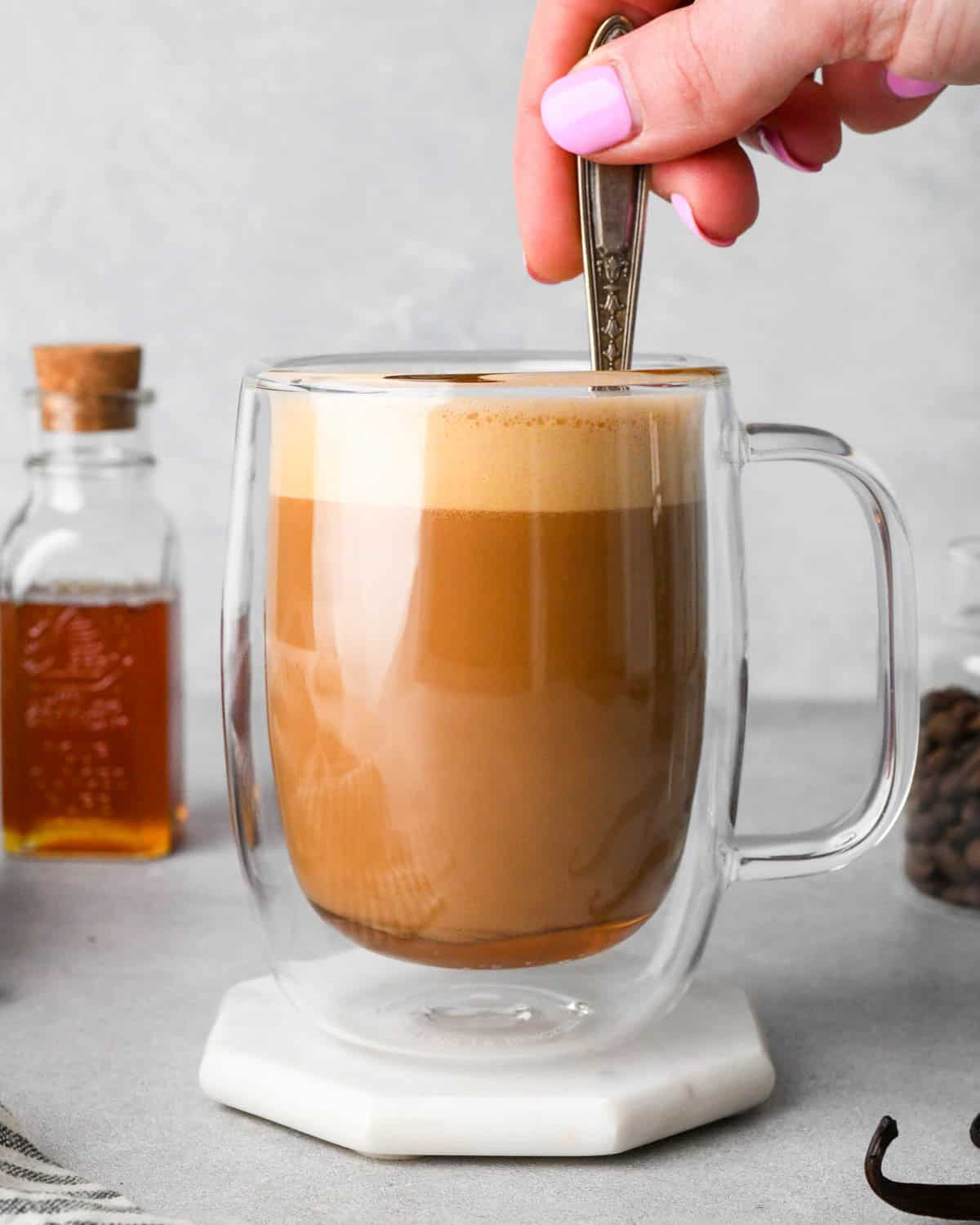a hand stirring a vanilla latte in a glass mug with a layer of foam on top. There's a jar of coffee beans in the background to the right, and a jar of honey in the backgroudn to the left.