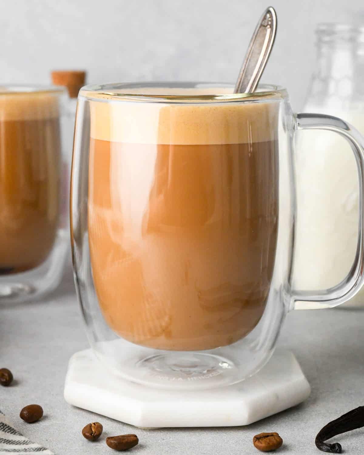 vanilla latte in a glass mug with a layer of foam on top and a silve spoon in it. There's another latte to the left of it and a bottle of milk in the background on the right