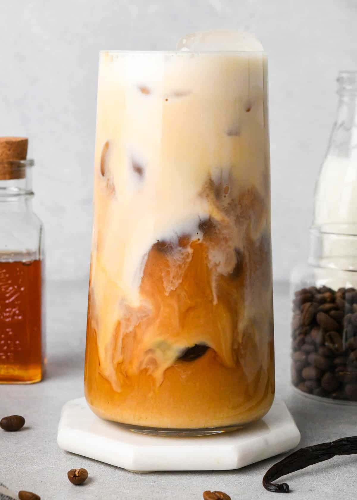 iced vanilla latte in a glass with the frothed milk giving it a two-tone look. surrounded by a jar of honey, coffee beans and milk