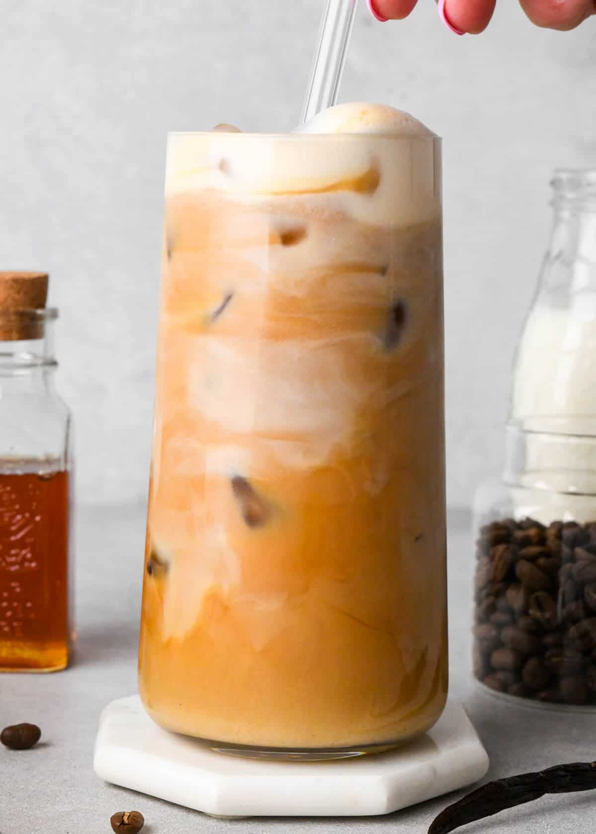 front view of a creamy iced vanilla latte being stirred so you can see swirls of milk and coffee in a glass. Surrounded by honey, coffee beans and milk all in jars