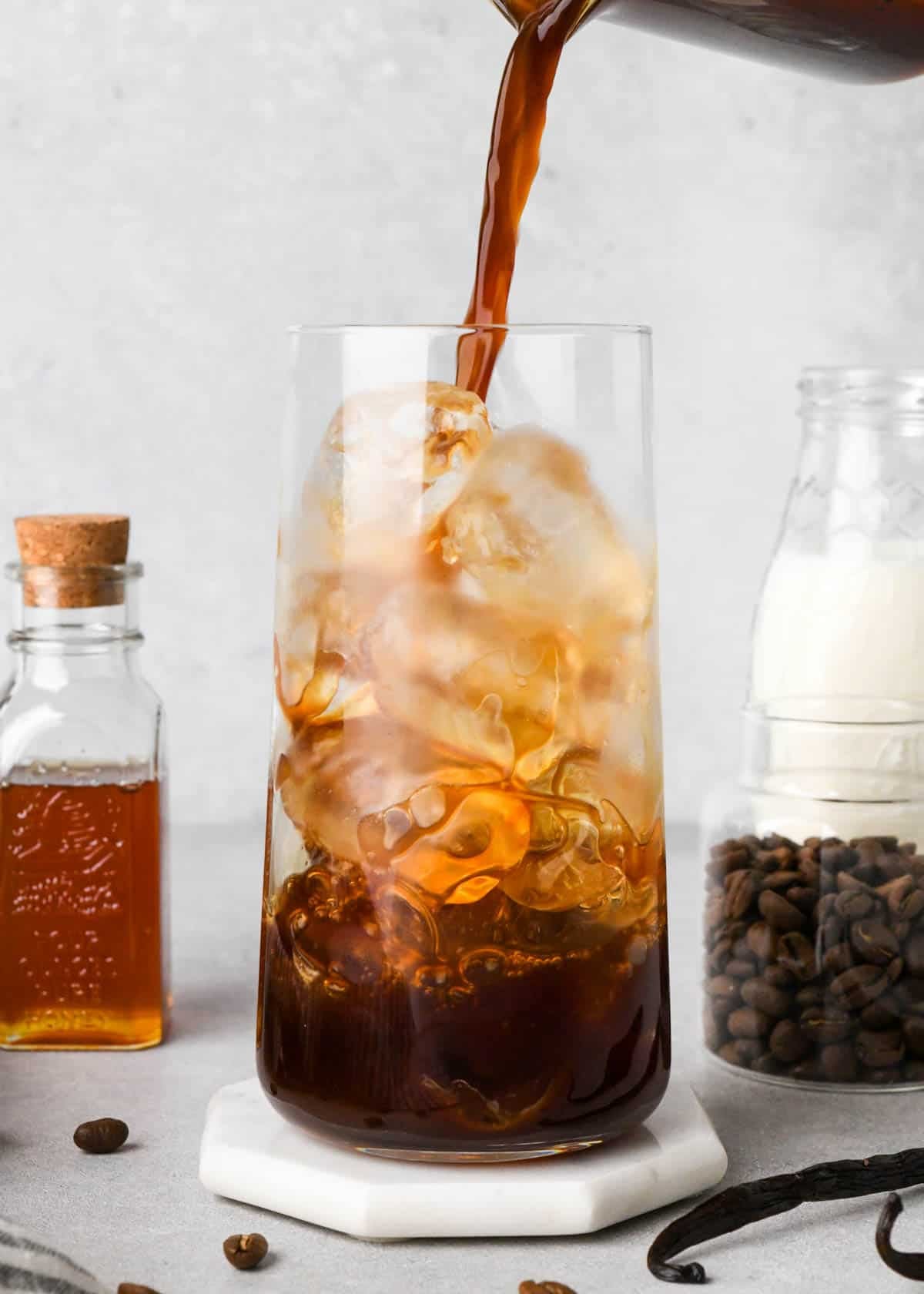 how to make an iced vanilla latte - brewed espresso being poured into a glass of ice with honey, coffee beans and milk in the backgrounds and vanilla beans and coffee beans in the foreground