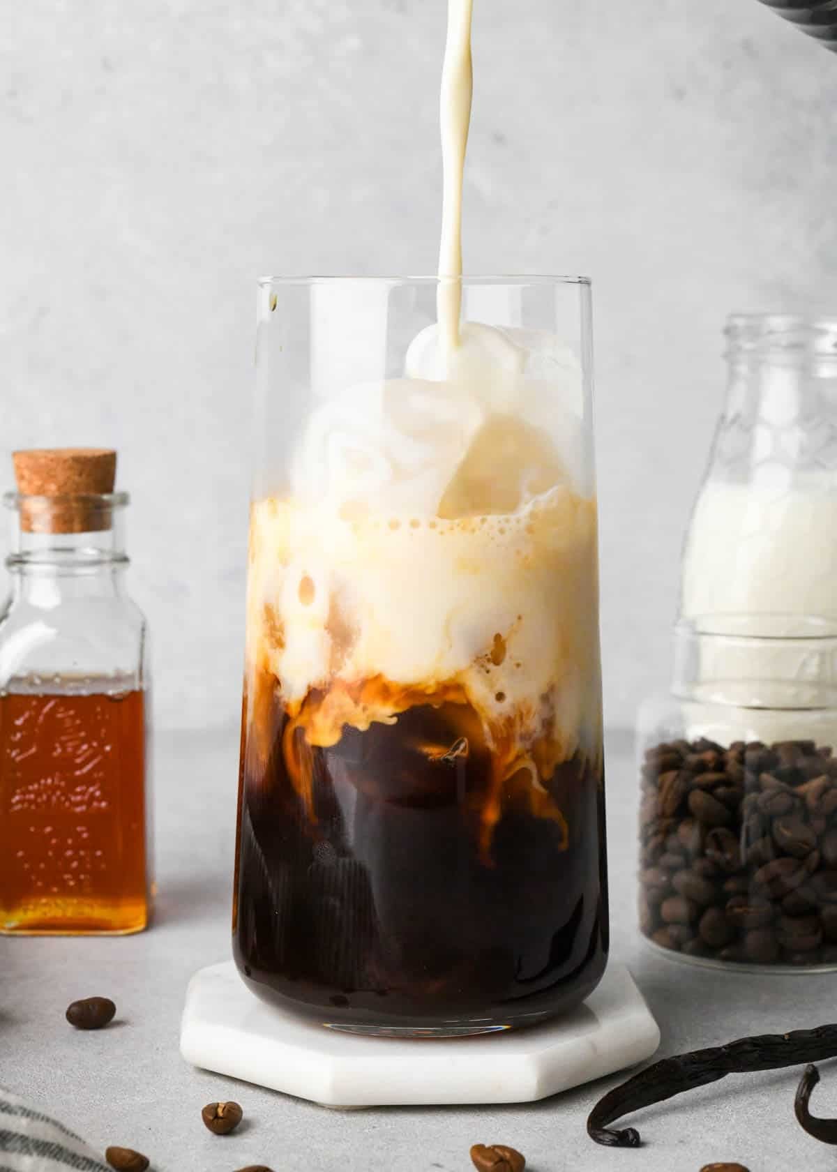 frothed milk being poured into an iced vanilla latte in a glass before it mixes in so the espresso and milk are two distinct layers, surrounded by a jar of honey, coffee beans and milk