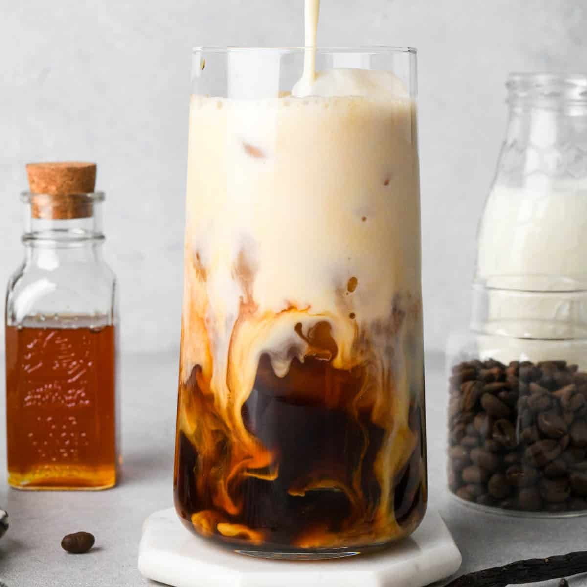 frothed milk being poured into an iced vanilla latte in a glass - the frothed milk is giving it a two-tone look. surrounded by a jar of honey, coffee beans and milk