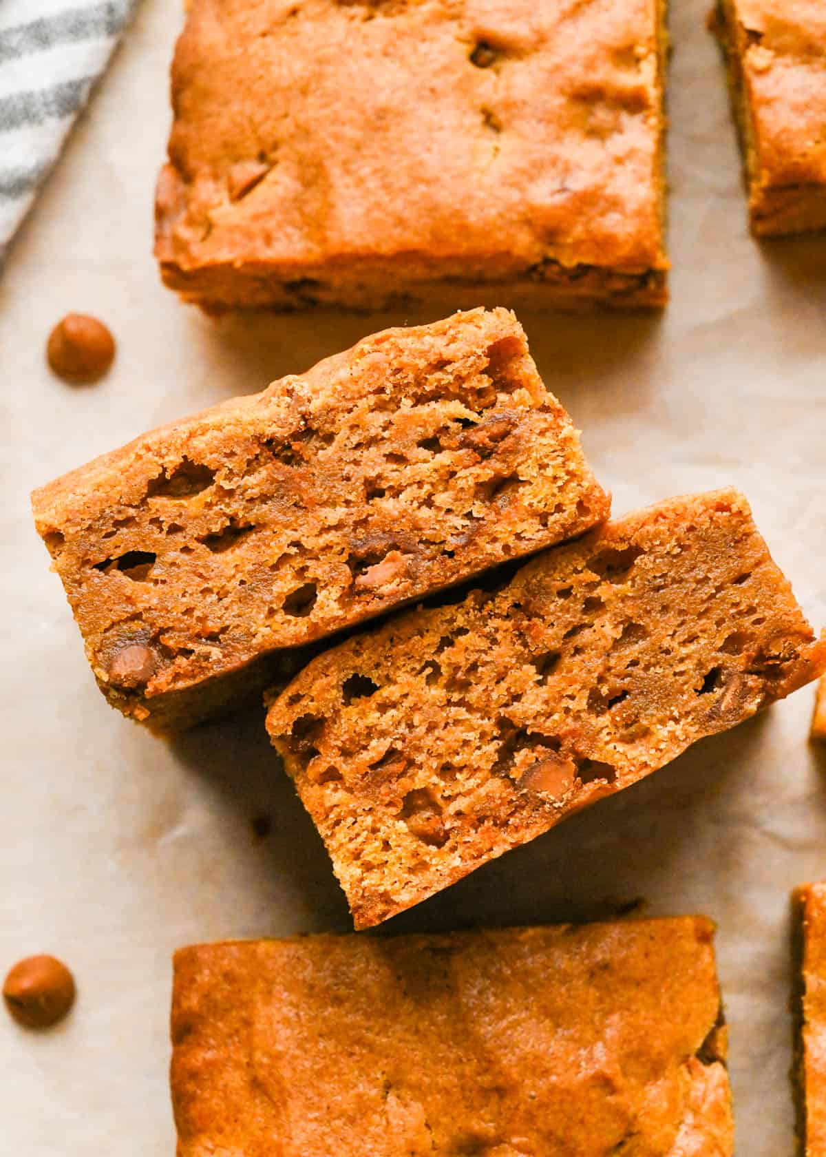 2 pumpkin blondies with cinnamon chips on their side so the chewy texture is showing on parchment paper surrounded by more pumpkin blondies and cinnamon chips