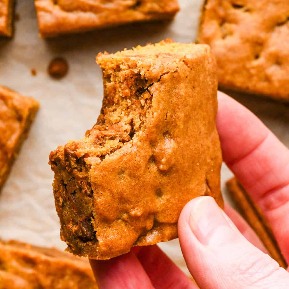 a hand holding a pumpkin blondie with a bite taken out of it so the chewy texture is visible. It's being held over parchment paper and more blondies