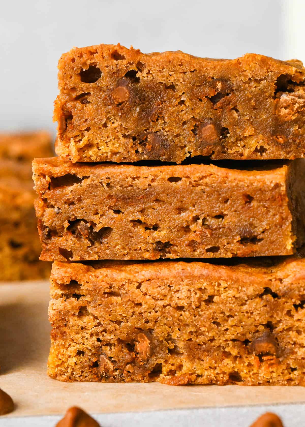 close up photo of a stack of 3 pumpkin blondies so the chewy, gooey texture and cinnamon chips inside are visible. The stack is on parchment paper and there are more blondies in the background