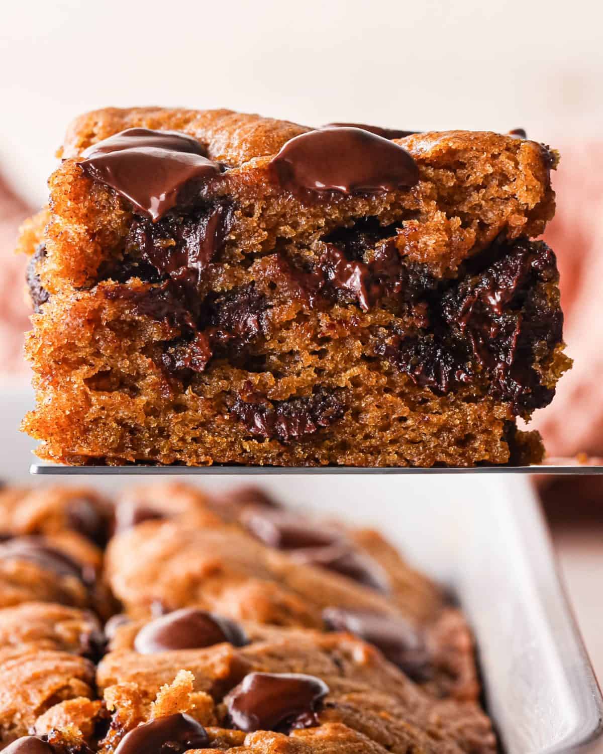 Close-up of a gooey pumpkin chocolate chip bar with a moist, spiced pumpkin crumb and abundant melted chocolate chips, with additional pumpkin chocolate chip bars blurred in the background.