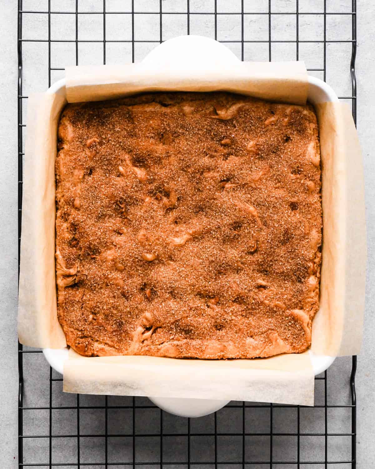 baked snickerdoodle blondies in a white ceramic, square, parchment-lined baking dish on a black wire cooling rack
