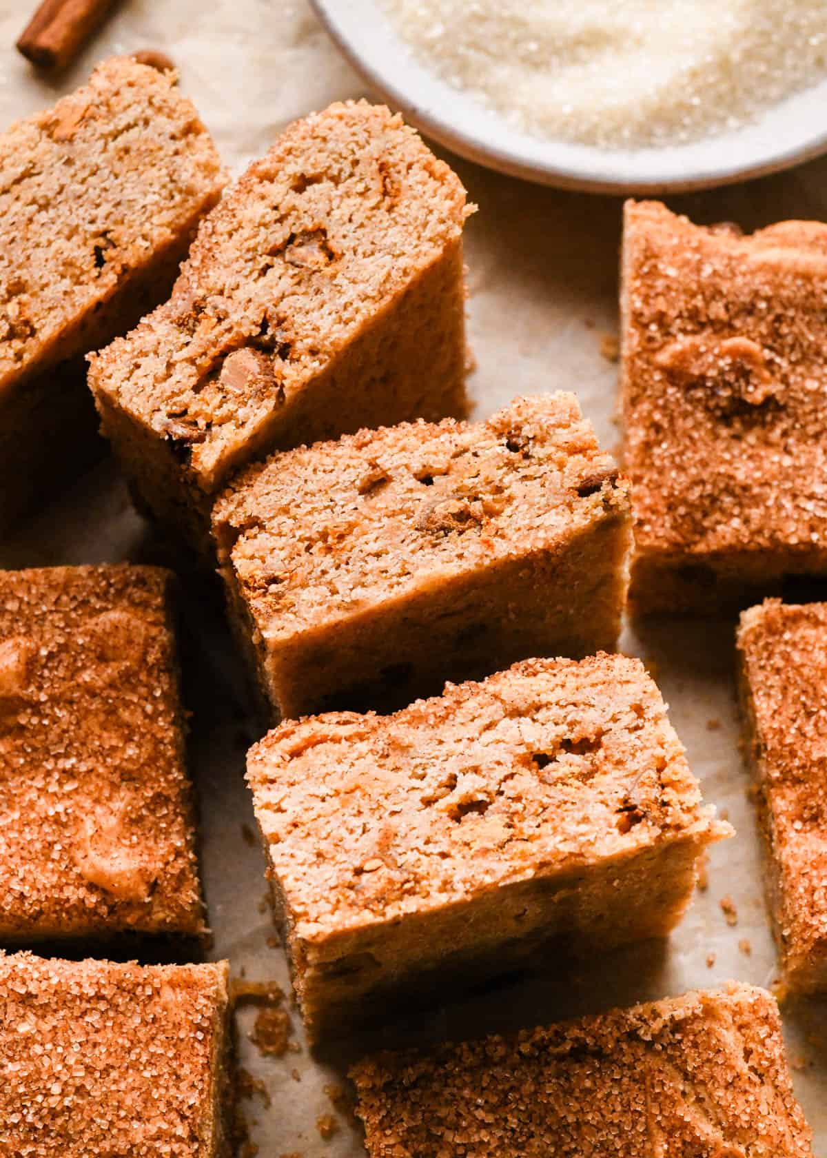 4 snickerdoodle blondies on their side so the chewy texture is showing, surrounded by more blondies on parchment paper there's a bowl of sugar in the top right side