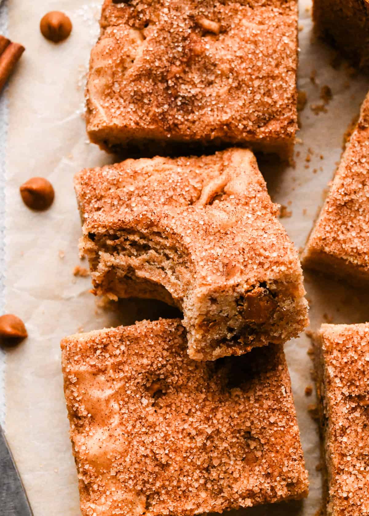 5 snickerdoodle blondies, one resting slightly on top of another one with a bite taken out of it so the chewy texture is visible on parchment paper surrounded by cinnamon chips